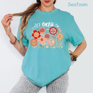 May include: A seafoam green short-sleeve t-shirt featuring a graphic print of colorful flowers with text that reads: it's OKAY to start over, make mistakes, be different, have bad days, and ask for help. The design includes various floral shapes.