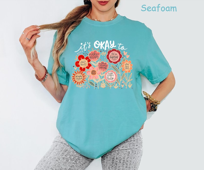 May include: A seafoam green short-sleeve t-shirt featuring a graphic print of colorful flowers with text that reads: it's OKAY to start over, make mistakes, be different, have bad days, and ask for help. The design includes various floral shapes.