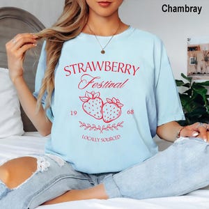 Op de afbeelding: Een lichtblauw t-shirt met rode tekst Strawberry Festival, 1968 en Locally Sourced, met een afbeelding van twee aardbeien en bladeren. Het t-shirt heeft een ronde hals en korte mouwen en wordt gedragen met een lichtblauwe spijkerbroek met scheuren.