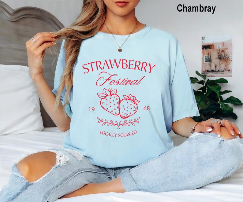 Op de afbeelding: Een lichtblauw t-shirt met rode tekst Strawberry Festival, 1968 en Locally Sourced, met een afbeelding van twee aardbeien en bladeren. Het t-shirt heeft een ronde hals en korte mouwen en wordt gedragen met een lichtblauwe spijkerbroek met scheuren.