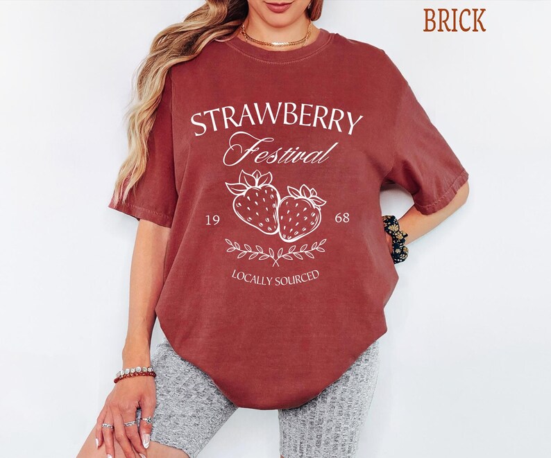 Op de afbeelding: Een baksteenrood t-shirt met korte mouwen en witte grafische tekst met de tekst Strawberry Festival, 1968 en Locally Sourced, samen met een illustratie van twee aardbeien en een bladerige tak in het midden van het kledingstuk.