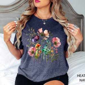 May include: Heather navy t-shirt featuring a watercolor floral design with pink, purple, orange, and green flowers. The shirt has a crew neck and short sleeves. The words "HEATHER NAVY" are printed in white at the bottom right.
