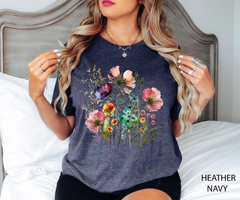 May include: Heather navy t-shirt featuring a watercolor floral design with pink, purple, orange, and green flowers. The shirt has a crew neck and short sleeves. The words "HEATHER NAVY" are printed in white at the bottom right.