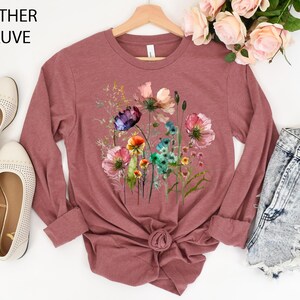 May include: Heather mauve long-sleeve shirt featuring a watercolor floral design. The shirt has a crew neck and a knotted hem. The text "Heather Mauve" is in the upper left corner. The shirt is paired with denim shorts and cream-colored flats.