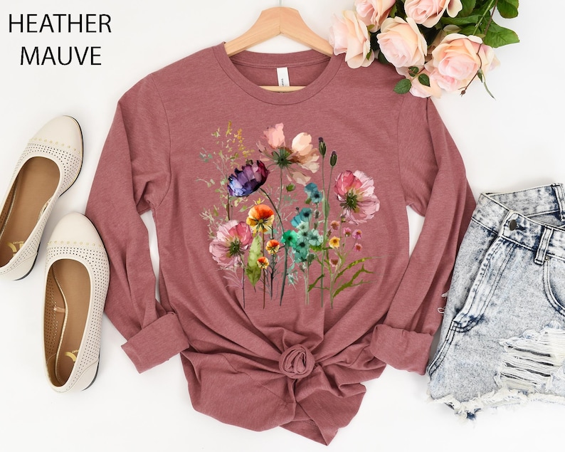 May include: Heather mauve long-sleeve shirt featuring a watercolor floral design. The shirt has a crew neck and a knotted hem. The text "Heather Mauve" is in the upper left corner. The shirt is paired with denim shorts and cream-colored flats.