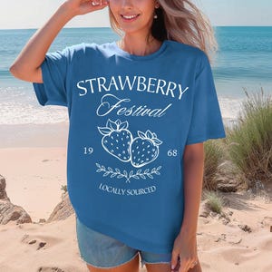 Op de afbeelding: Een blauw t-shirt met korte mouwen en een witte grafische opdruk op de voorzijde, bestaande uit de tekst STRAWBERRY Festival, de getallen 19 en 68, een illustratie van twee aardbeien met blaadjes en de tekst LOCALLY SOURCED.