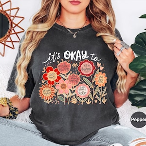 May include: A charcoal gray short-sleeve t-shirt featuring a graphic of colorful flowers with text that reads: it's OKAY to make mistakes, be different, start over, have strong emotions, have bad days, not know everything, ask for help.