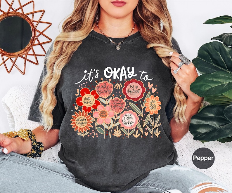 May include: A charcoal gray short-sleeve t-shirt featuring a graphic of colorful flowers with text that reads: it's OKAY to make mistakes, be different, start over, have strong emotions, have bad days, not know everything, ask for help.