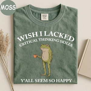 May include: A moss green crew neck t-shirt featuring a graphic of a standing frog holding a small orange mug. The text reads WISH I LACKED CRITICAL THINKING SKILLS above the frog and Y'ALL SEEM SO HAPPY below the frog in white lettering.