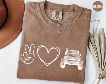 May include: Espresso-colored t-shirt with a white graphic of a peace sign, heart, and off-road vehicle. The shirt has a crew neck and short sleeves. A silver necklace with a pendant is draped across the shirt.