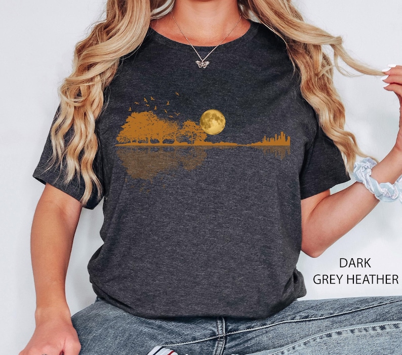 Pode incluir: T-shirt cinza escuro mesclada com um design de paisagem. Apresenta uma lua cheia, &aacute;rvores e p&aacute;ssaros em voo, em tons de ouro e marrom. O design reflete-se abaixo. O texto "DARK GREY HEATHER" &eacute; vis&iacute;vel.