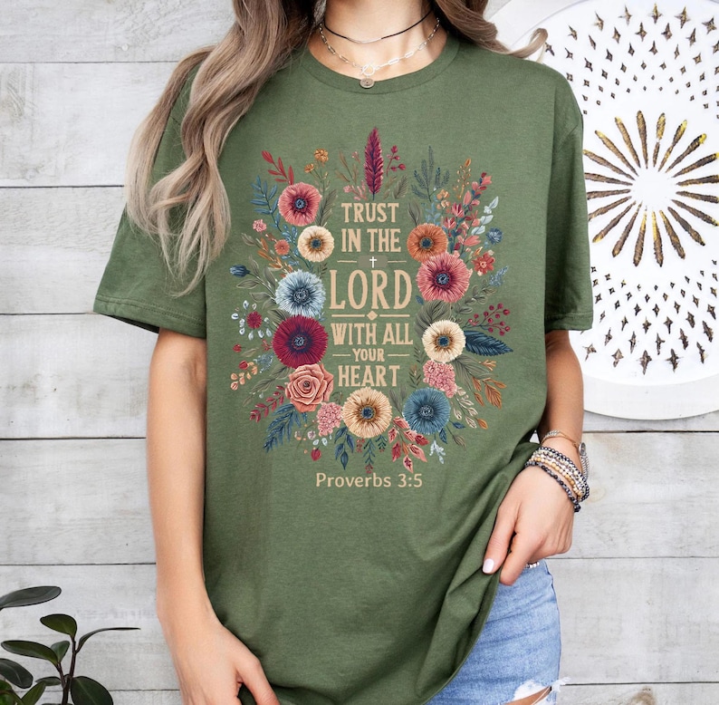 May include: Olive green t-shirt featuring a floral design with the text "Trust in the Lord with all your heart" and "Proverbs 3:5". The design includes colorful flowers and foliage.