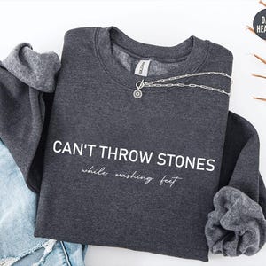 May include: A dark heather grey crewneck sweatshirt featuring the white text CAN'T THROW STONES while washing feet printed on the front. The garment has long sleeves with ribbed cuffs and a silver-toned toggle necklace resting on the neckline.