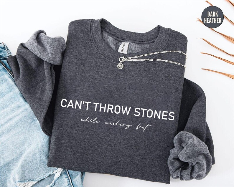 May include: A dark heather grey crewneck sweatshirt featuring the white text CAN'T THROW STONES while washing feet printed on the front. The garment has long sleeves with ribbed cuffs and a silver-toned toggle necklace resting on the neckline.