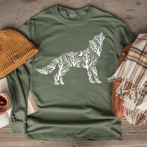 May include: An olive green long-sleeve shirt with a white wolf design made of tree branches and a crescent moon. A mustard yellow beanie, a plaid scarf, and a leather bag are also in the image.