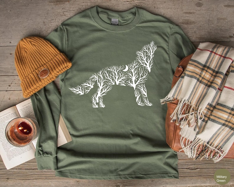 May include: An olive green long-sleeve shirt with a white wolf design made of tree branches and a crescent moon. A mustard yellow beanie, a plaid scarf, and a leather bag are also in the image.