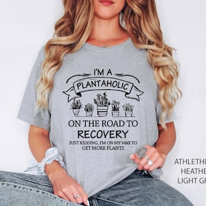 May include: A light grey short-sleeve t-shirt featuring black text that reads I'M A PLANTAHOLIC ON THE ROAD TO RECOVERY JUST KIDDING, I'M ON MY WAY TO GET MORE PLANTS, accompanied by a graphic illustration of five potted plants in a row.