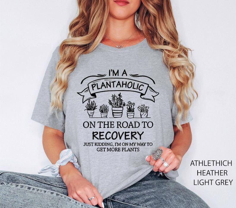 May include: A light grey short-sleeve t-shirt featuring black text that reads I'M A PLANTAHOLIC ON THE ROAD TO RECOVERY JUST KIDDING, I'M ON MY WAY TO GET MORE PLANTS, accompanied by a graphic illustration of five potted plants in a row.