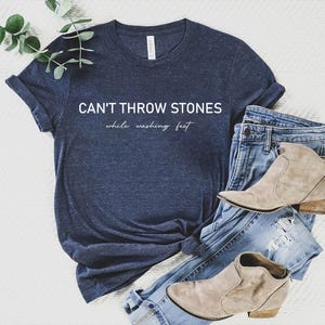 May include: A heather midnight navy short-sleeve t-shirt with the white text CAN'T THROW STONES while washing feet printed on the front. The shirt is paired with light blue distressed denim jeans and two beige suede ankle boots with low heels.