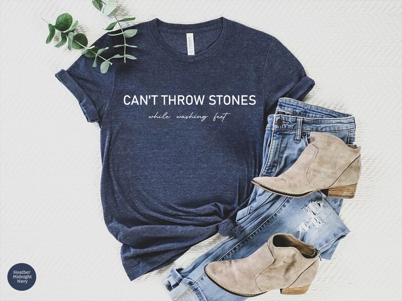 May include: A heather midnight navy short-sleeve t-shirt with the white text CAN'T THROW STONES while washing feet printed on the front. The shirt is paired with light blue distressed denim jeans and two beige suede ankle boots with low heels.