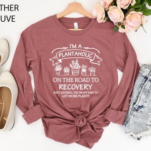 May include: A heather mauve long-sleeve shirt featuring white text that reads I'M A PLANTAHOLIC ON THE ROAD TO RECOVERY JUST KIDDING, I'M ON MY WAY TO GET MORE PLANTS, accompanied by a graphic illustration of five potted plants in a row.