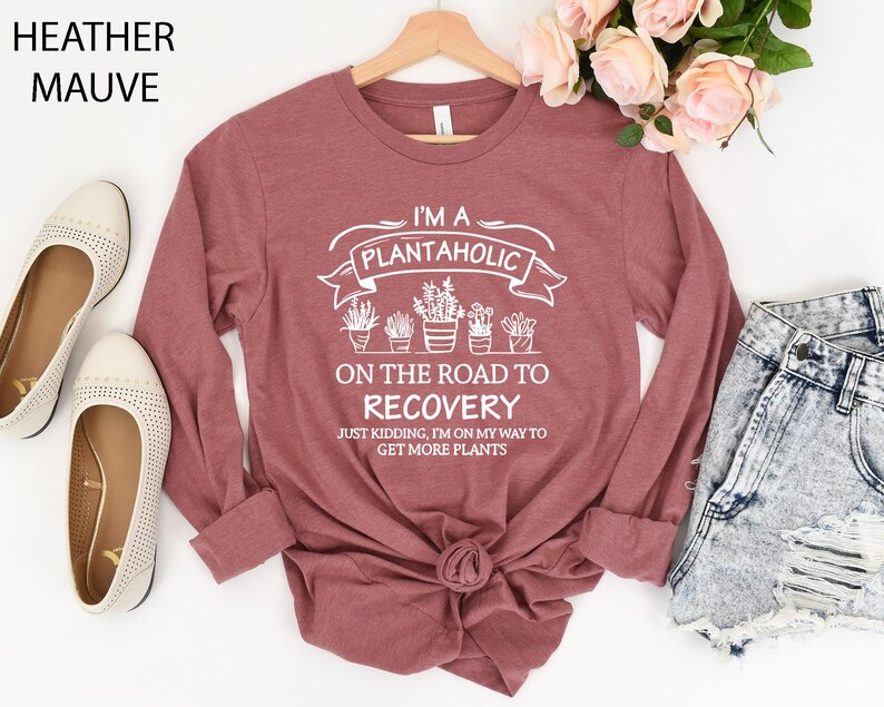May include: A heather mauve long-sleeve shirt featuring white text that reads I'M A PLANTAHOLIC ON THE ROAD TO RECOVERY JUST KIDDING, I'M ON MY WAY TO GET MORE PLANTS, accompanied by a graphic illustration of five potted plants in a row.