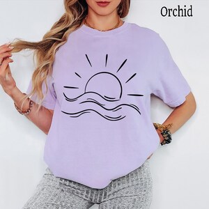 Pu&ograve; includere: Una t-shirt a maniche corte color orchidea con una grafica a linee nere che raffigura un sole che sorge sopra le onde dell'oceano. La parola Orchid &egrave; stampata nell'angolo in alto a destra. La maglietta ha un girocollo e una vestibilit&agrave; ampia.