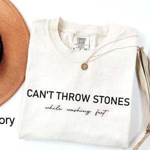 K&ouml;nnte beinhalten: Ein T-Shirt in Elfenbein mit dem in Schwarz gedruckten Text "CAN'T THROW STONES while washing feet".