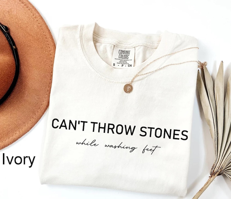 K&ouml;nnte beinhalten: Ein T-Shirt in Elfenbein mit dem in Schwarz gedruckten Text "CAN'T THROW STONES while washing feet".