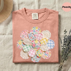 May include: Peach-colored t-shirt with a floral design made of various patterned fabric circles. The flower design includes pink, blue, yellow, and white fabrics. The shirt has a Comfort Colors tag.