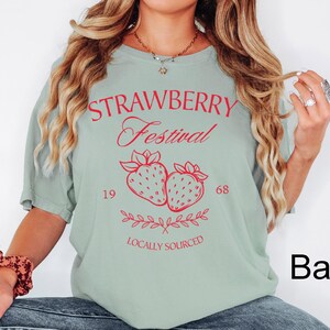 Op de afbeelding: Een saliegroen t-shirt met korte mouwen en rode tekst met de woorden STRAWBERRY Festival, 1968 en LOCALLY SOURCED. De afbeelding bevat twee ge&iuml;llustreerde aardbeien en een takje met blaadjes in het midden van het kledingstuk.