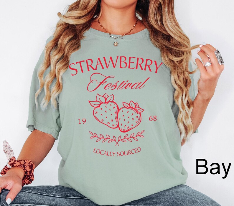 Op de afbeelding: Een saliegroen t-shirt met korte mouwen en rode tekst met de woorden STRAWBERRY Festival, 1968 en LOCALLY SOURCED. De afbeelding bevat twee ge&iuml;llustreerde aardbeien en een takje met blaadjes in het midden van het kledingstuk.