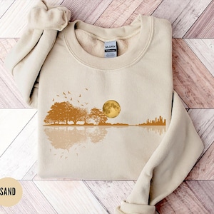 Pode incluir: Um moletom cor de areia com um design de paisagem. O design inclui &aacute;rvores, uma lua cheia, p&aacute;ssaros e um horizonte urbano refletido na &aacute;gua. O moletom &eacute; feito de um material macio.