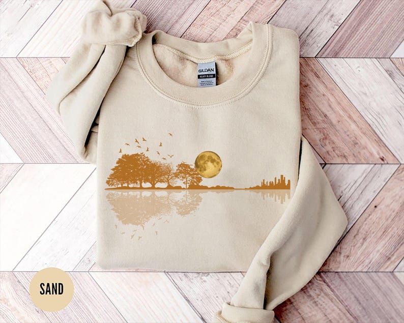 Pode incluir: Um moletom cor de areia com um design de paisagem. O design inclui &aacute;rvores, uma lua cheia, p&aacute;ssaros e um horizonte urbano refletido na &aacute;gua. O moletom &eacute; feito de um material macio.
