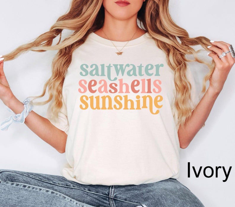 K&ouml;nnte beinhalten: Elfenbeinfarbenes T-Shirt mit dem Schriftzug "saltwater seashells sunshine" in Pastellfarben. Das Shirt hat einen Rundhalsausschnitt und eine lockere Passform. Der Text ist in einer verspielten Retro-Schriftart.