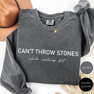 K&ouml;nnte beinhalten: Ein dunkelgrauer Pullover mit dem Text "CAN'T THROW STONES while washing feet" in wei&szlig;en Buchstaben. Der Pullover hat einen Used-Look und ist von Comfort Colors.