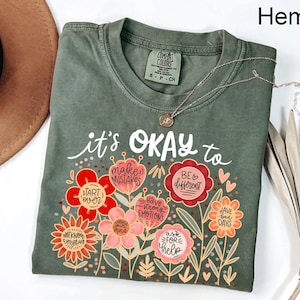 May include: A hemp-colored t-shirt featuring a graphic of colorful flowers with text that reads: it's OKAY to start over, make mistakes, be different, have strong emotions, change your mind, ask for help, have bad days, and not know everything.