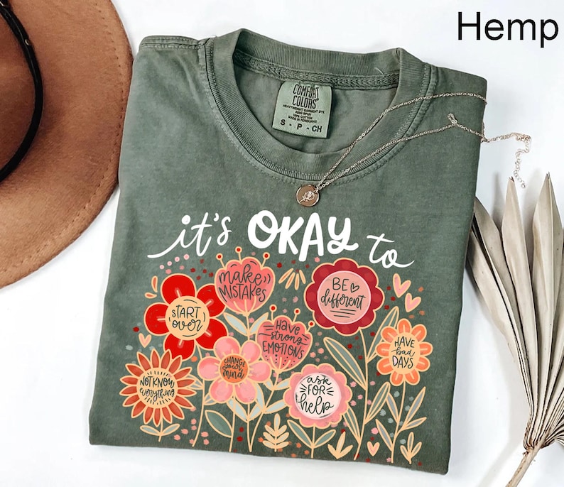 May include: A hemp-colored t-shirt featuring a graphic of colorful flowers with text that reads: it's OKAY to start over, make mistakes, be different, have strong emotions, change your mind, ask for help, have bad days, and not know everything.