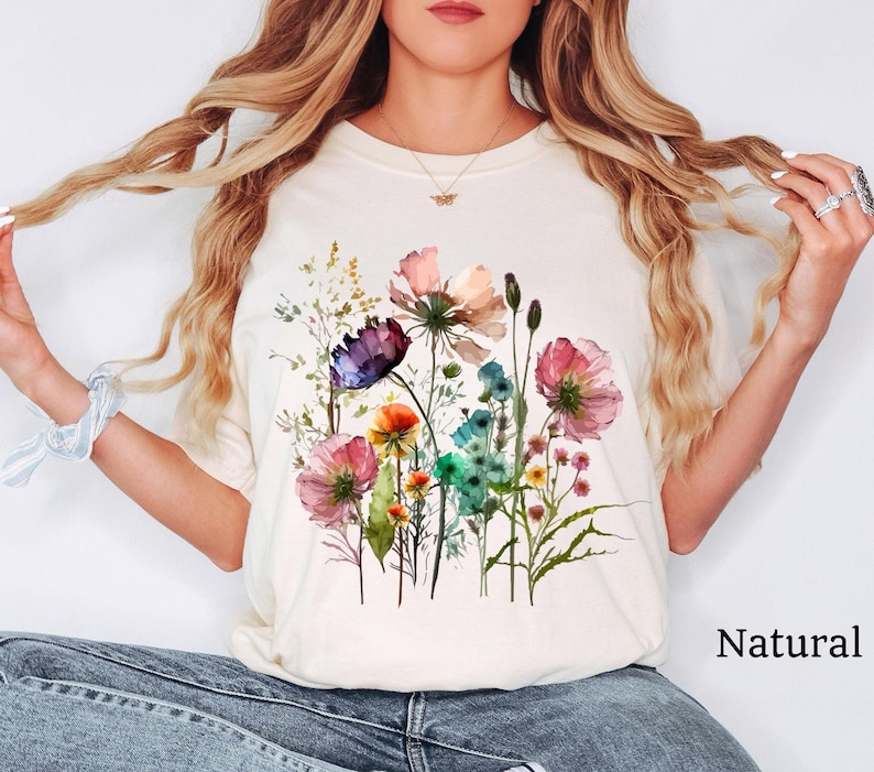 May include: A white t-shirt with a watercolor floral print featuring pink, purple, blue, and yellow flowers. The text "Natural" is printed on the bottom right corner of the image.