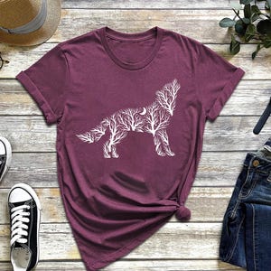 May include: A maroon t-shirt featuring a white graphic of a wolf silhouette composed of tree branches and a crescent moon. The shirt is styled with blue jeans, black sneakers, and a straw hat.