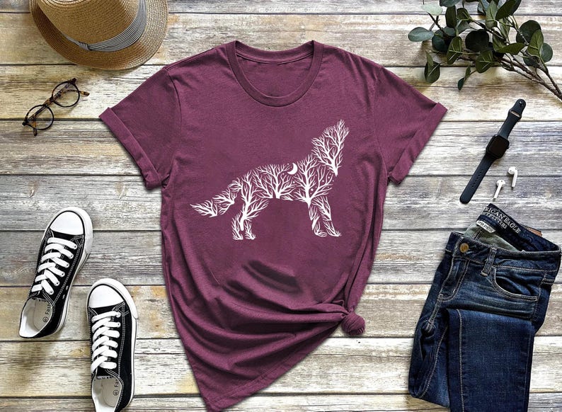 May include: A maroon t-shirt featuring a white graphic of a wolf silhouette composed of tree branches and a crescent moon. The shirt is styled with blue jeans, black sneakers, and a straw hat.