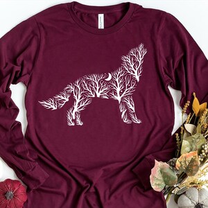 May include: Maroon long-sleeve shirt featuring a white wolf design. The wolf is created from a tree branch pattern with a crescent moon. The shirt is a comfortable, casual style.