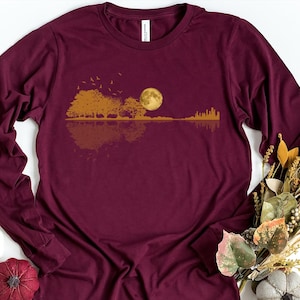 Pode incluir: Camiseta de manga comprida bord&ocirc; com um design de paisagem dourada. O gr&aacute;fico inclui &aacute;rvores, uma lua cheia e um horizonte urbano refletido na &aacute;gua. A camiseta &eacute; de cor bord&ocirc; lisa.