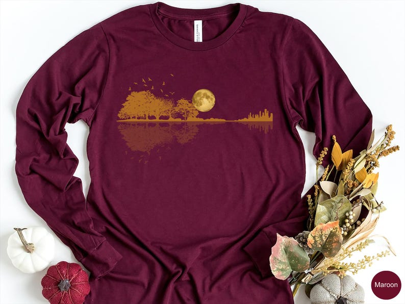 Pode incluir: Camiseta de manga comprida bord&ocirc; com um design de paisagem dourada. O gr&aacute;fico inclui &aacute;rvores, uma lua cheia e um horizonte urbano refletido na &aacute;gua. A camiseta &eacute; de cor bord&ocirc; lisa.