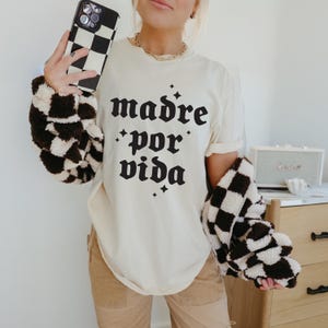 May include: A white t-shirt with black text that reads "madre por vida" in a gothic font. The shirt is being worn by a person with a black and white checkered jacket.
