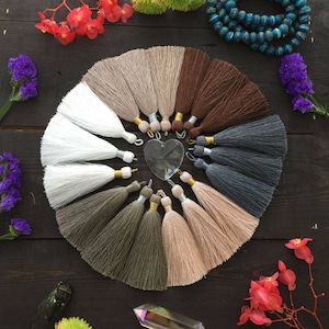 Könnte beinhalten: Eine Sammlung von 10 verschiedenen farbigen Quasten, die in einem Kreis auf einer dunklen Holzoberfläche angeordnet sind. Die Quasten bestehen aus weichem, flauschigem Garn und haben einen goldenen Metallring oben. Die Quasten sind alle in verschiedenen Farben, darunter Weiß, Braun, Grau und Beige.