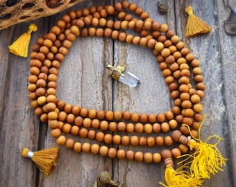 May include: A wooden mala necklace with brown beads and yellow tassels. The necklace is laid out on a wooden surface with a piece of desert wood and other decorative items.