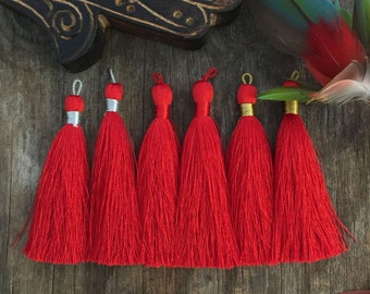 May include: Six bright red tassels with silver and gold accents are arranged on a rustic wooden surface. Decorative elements include a carved wooden hand, feathers, a crystal, and decorative arrows. The tassels are a vibrant accent.