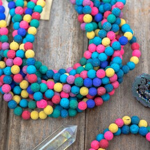 May include: A close-up of a pile of colorful lava rock beads. The beads are round and have a rough, textured surface. They are arranged in a circle, with the colors yellow, pink, blue, and green.
