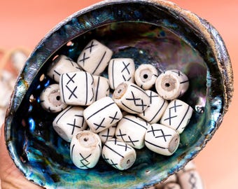 May include: Abalone shell bowl filled with white beads featuring black crosshatch designs. The beads are cylindrical with a hole through the center. The shell has iridescent blue, green, and purple hues. A hand holds the shell, with bracelets visible.